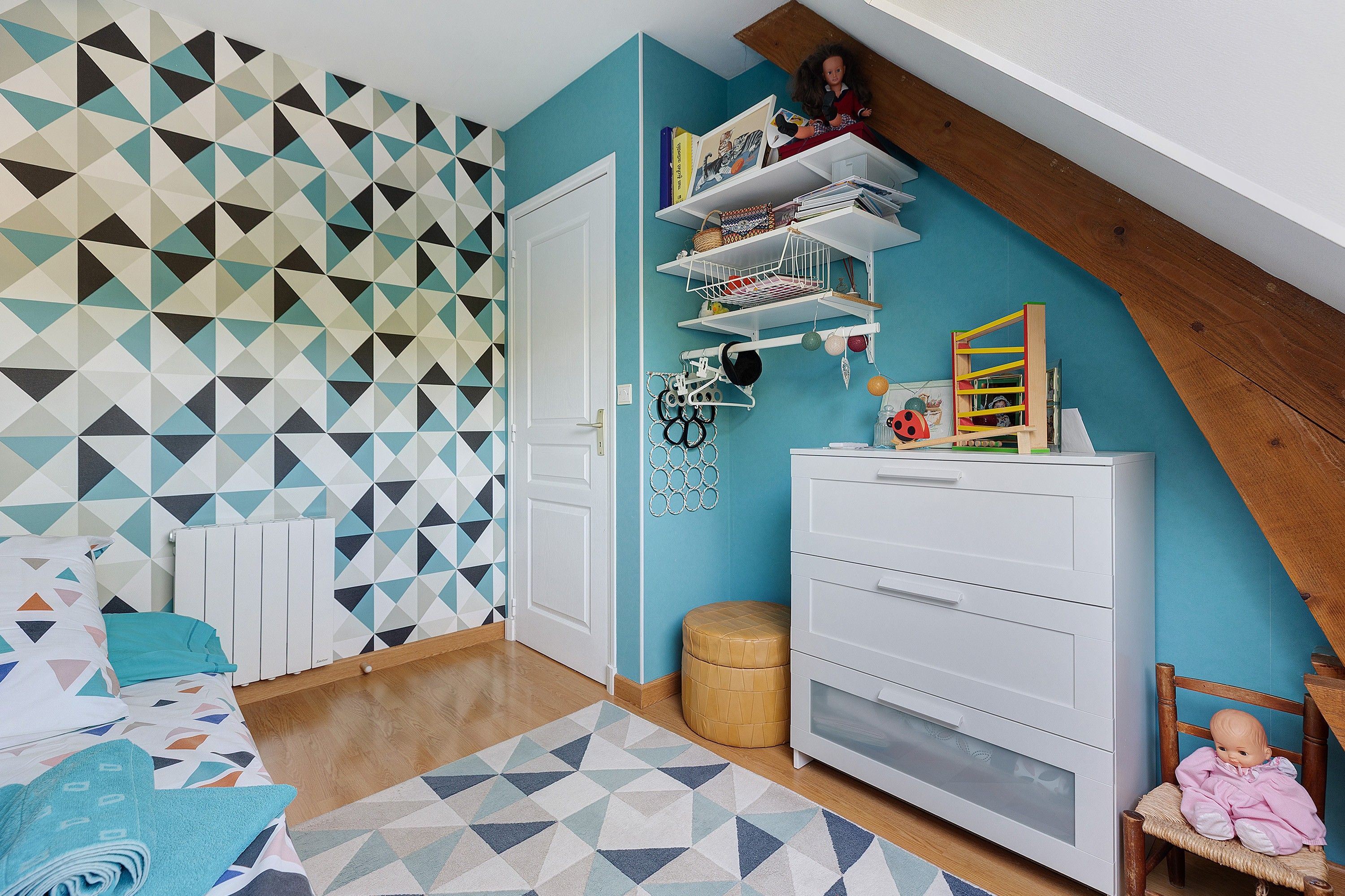 Une pièce avec un mur orné d’un motif géométrique, une commode blanche, une chaise en bois et une petite table sur laquelle est posée une poupée.