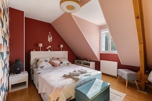A bedroom with a sloped ceiling, a bed with multiple pillows, bedside tables, a lamp, a radiator, and a window.
