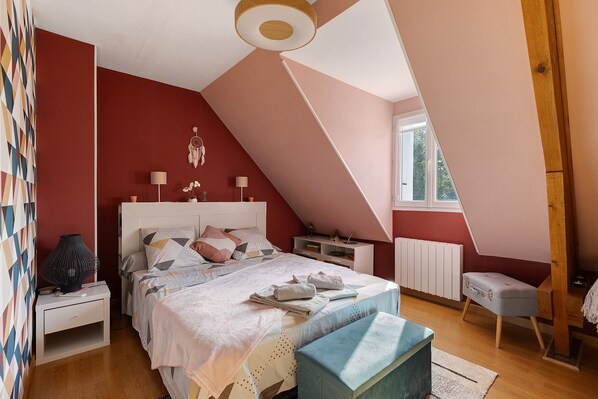 A bedroom with a sloped ceiling, a bed with multiple pillows, bedside tables, a lamp, a radiator, and a window.