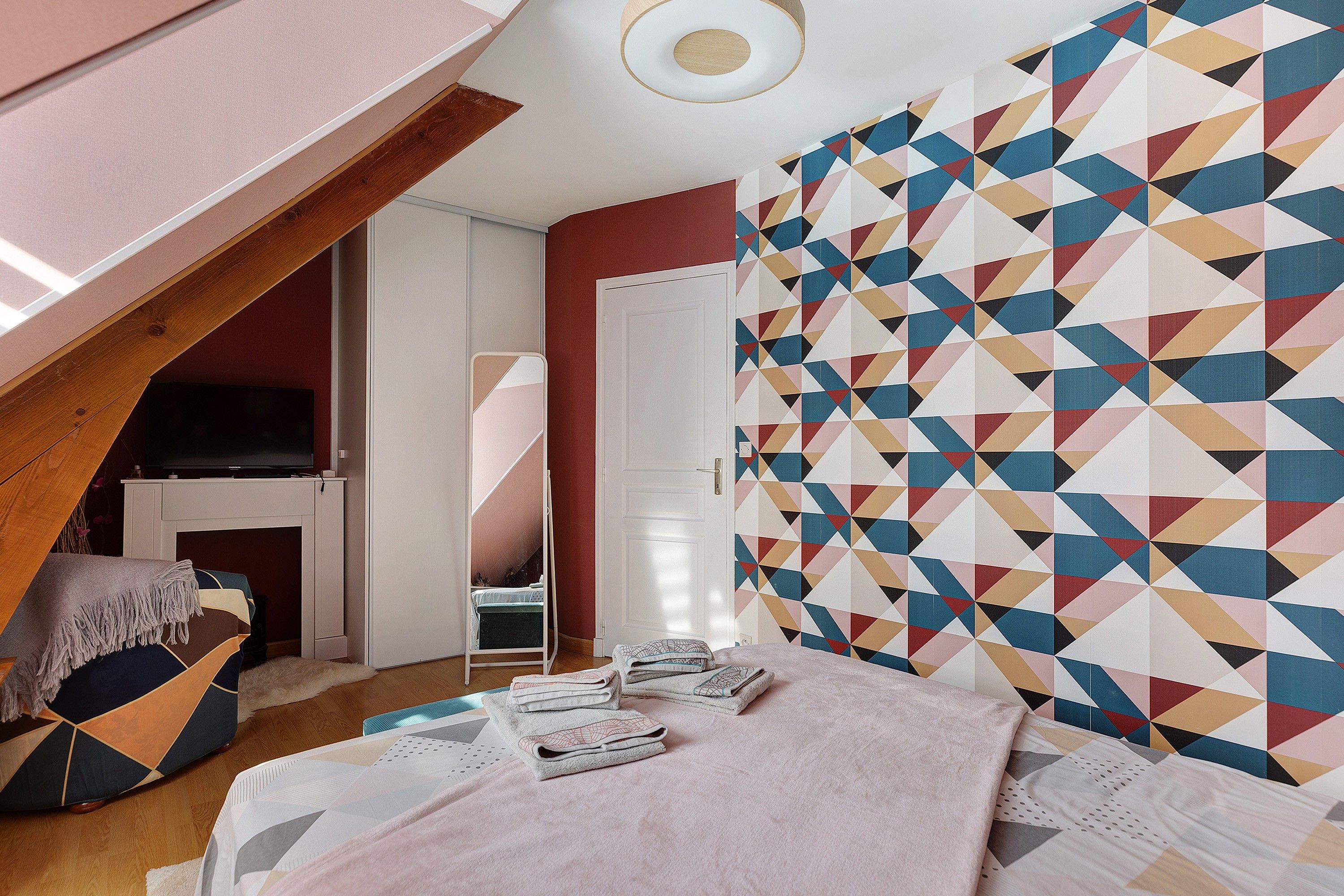 Un dormitorio con techo inclinado, una cama con almohadas y una manta, un televisor y una pared con estampado geométrico.