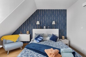 A bedroom with a sloped ceiling, a bed with patterned bedding, a bedside table with lamps, and a chair with a yellow throw.