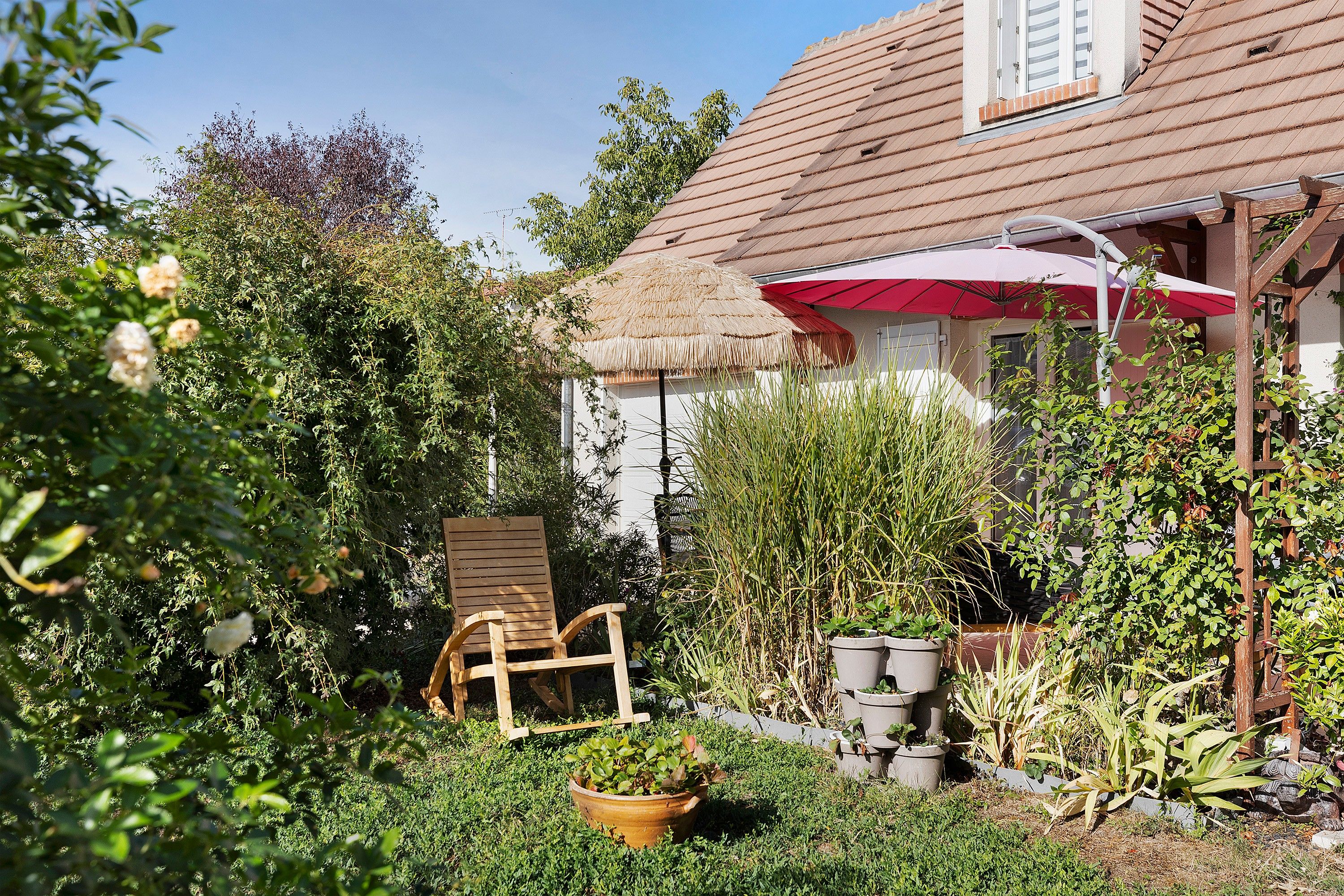 Un jardin avec une chaise en bois, des plantes en pot et une structure dotée d’un toit de chaume.