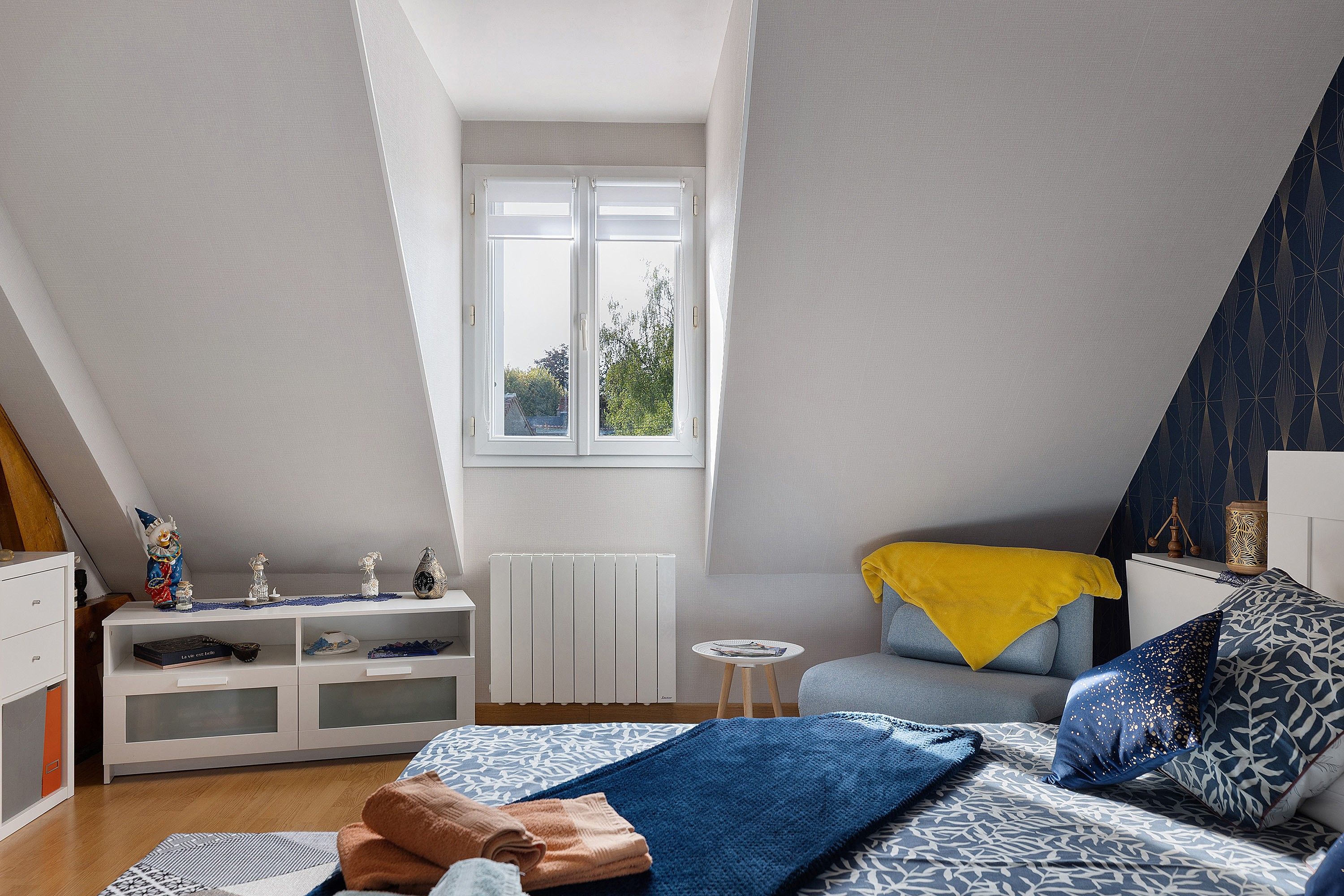Un dormitorio con techo inclinado, una ventana, una cama con ropa de cama estampada, una mesita de noche y un pequeño aparador blanco.