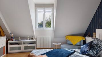 Un dormitorio con techo inclinado, una ventana, una cama con ropa de cama estampada, una mesita de noche y un pequeño aparador blanco.