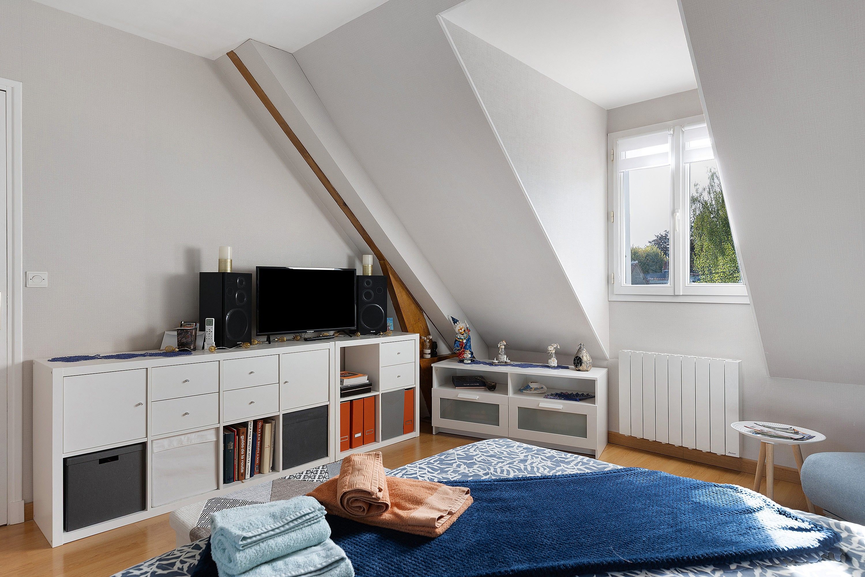 Quarto moderno com teto inclinado, TV sobre um armário branco, radiador e janela com vista para área verde.