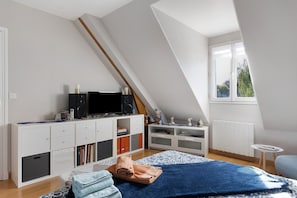 A modern bedroom with a sloped ceiling, a TV on a white cabinet, a radiator, and a window with a view of greenery.