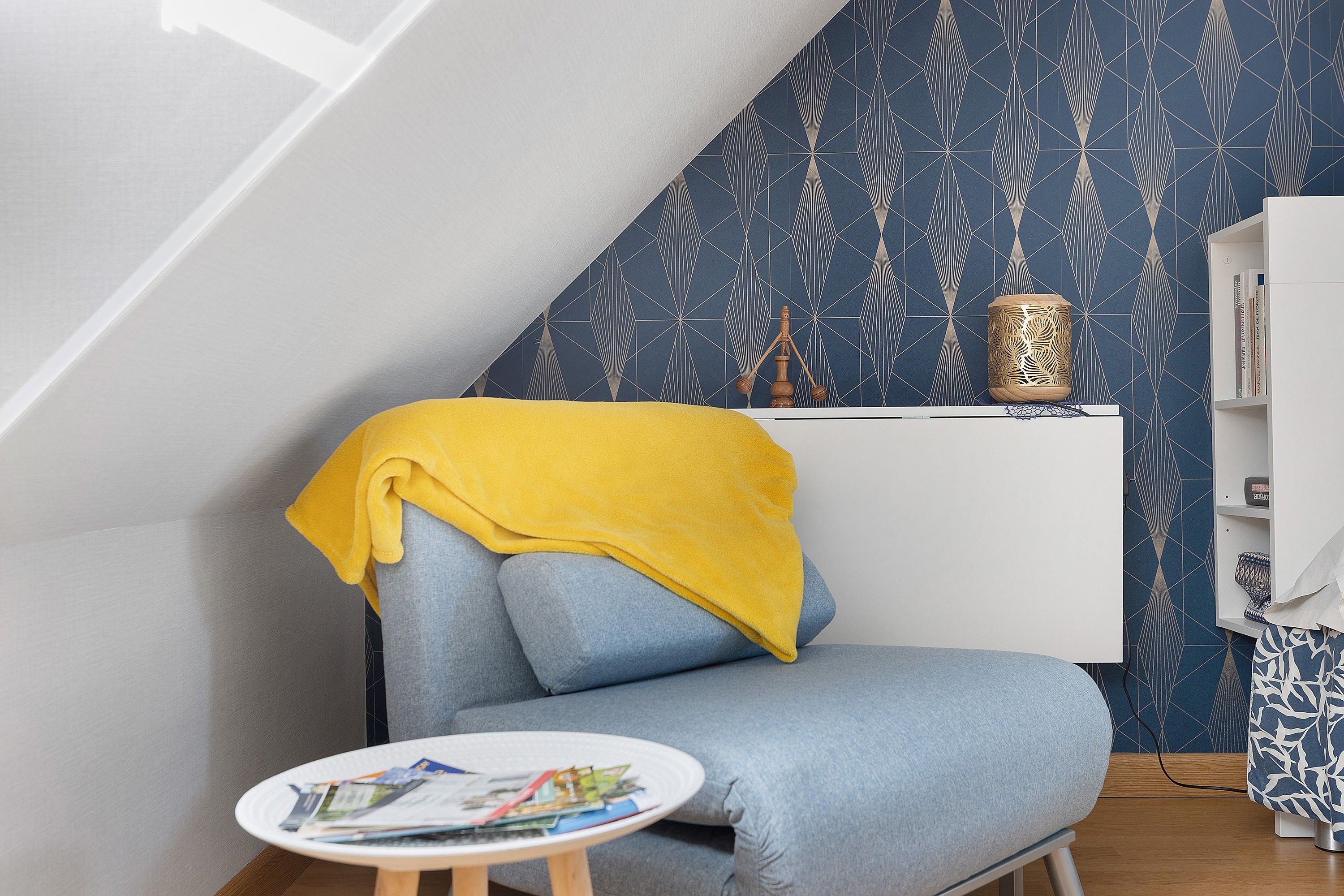 Un coin de pièce avec un mur bleu à motifs, une bibliothèque blanche et une petite table ronde sur laquelle sont posés des magazines.