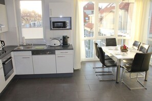 A compact kitchen with a microwave, toaster, and coffee maker, a dining area with a table and chairs, and a view of buildings through large windows. - Holiday apartment with 2 bedrooms (Großenbrode)