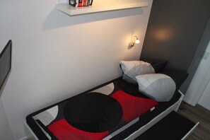 A small, single-bed room with a bedside table, a wall-mounted shelf with books, and a wall-mounted light fixture. - Holiday apartment with 2 bedrooms (Großenbrode)