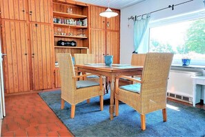 A dining area with a wooden table and wicker chairs, a bookshelf with various items, and a window with a curtain. - Holiday home in Schlei with 2 bedrooms (Brodersby-Burg)
