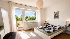 A modern bedroom with a bed, a desk, a chair, a radiator, and a window with a view of greenery. - Pulheim Living 2 (Pulheim)