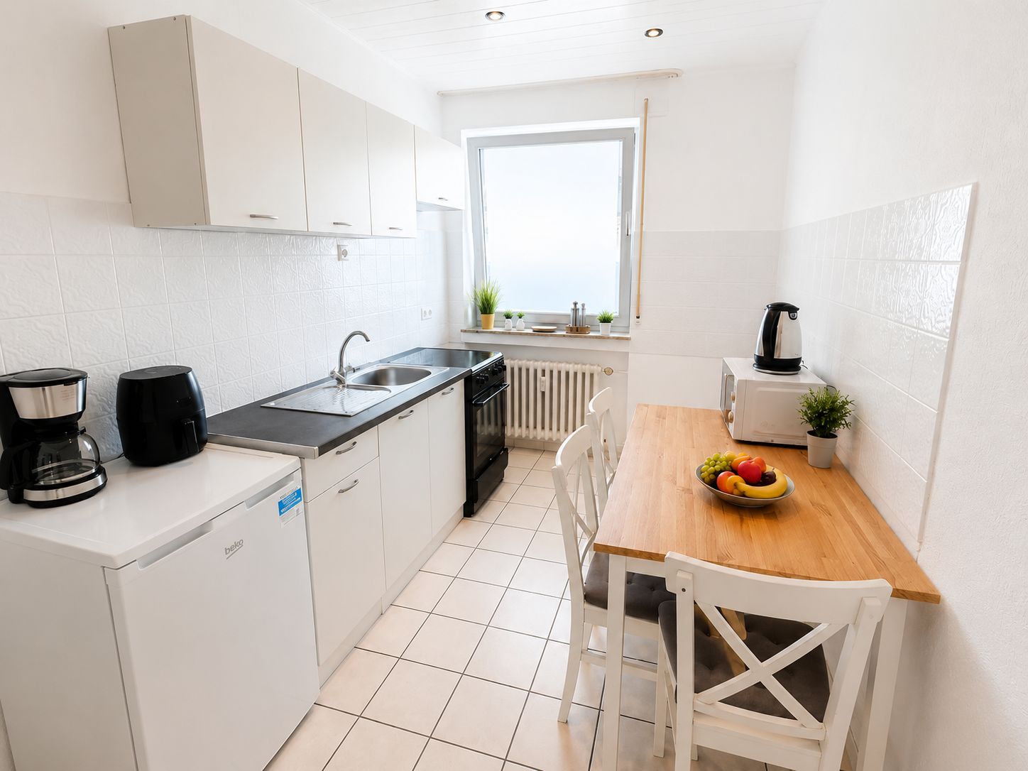 Una cocina compacta con armarios blancos, una estufa negra, un fregadero y una mesa de comedor de madera con un bol de fruta.