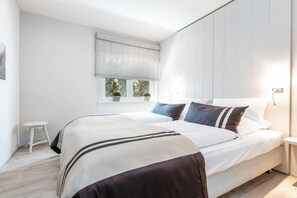 A bedroom with a bed, bedside tables, a window with blinds, and a small stool. - Holiday home with 2 bedrooms (Heiligenhafen)