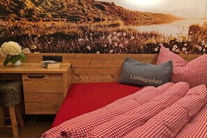 Ein Schlafzimmer mit einem hölzernen Kopfteil, einer roten Bettdecke, karierten Kissen, einem kleinen Nachttisch mit einem Buch und einer Blumenvase sowie einer Wandmalerei von einer Landschaft.