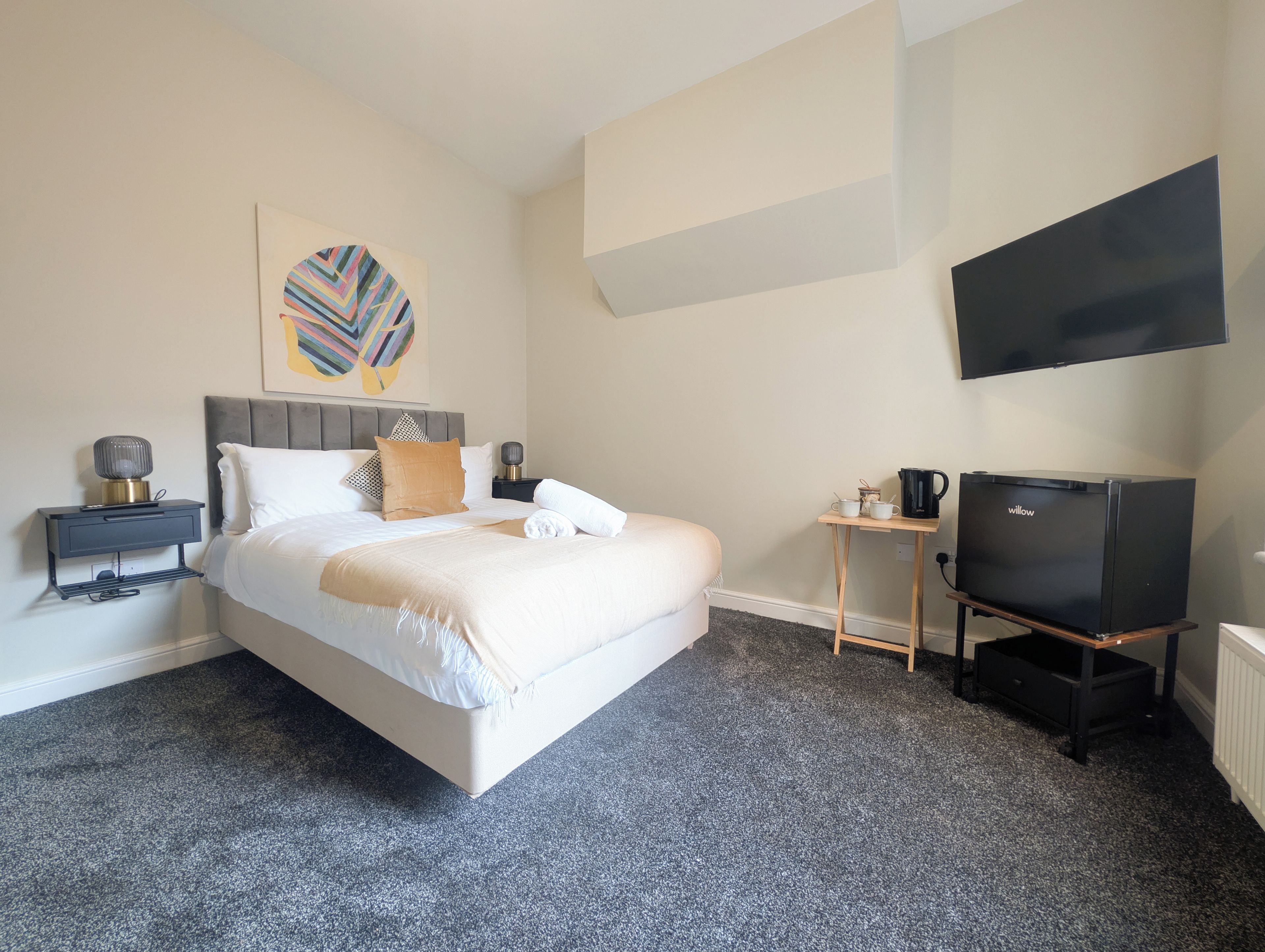 A hotel room with a bed, a bedside table with a lamp, a small table with a coffee maker, and a TV on a stand.