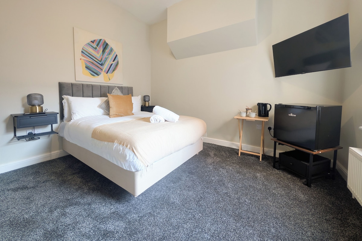 Ett hotellrum med en säng, ett sängbord med en lampa, ett litet bord med en kaffebryggare och en tv som står på ett stativ.