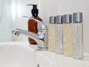 Un baño con un lavamanos blanco, un grifo plateado y cuatro botellas de champú y gel de ducha.
