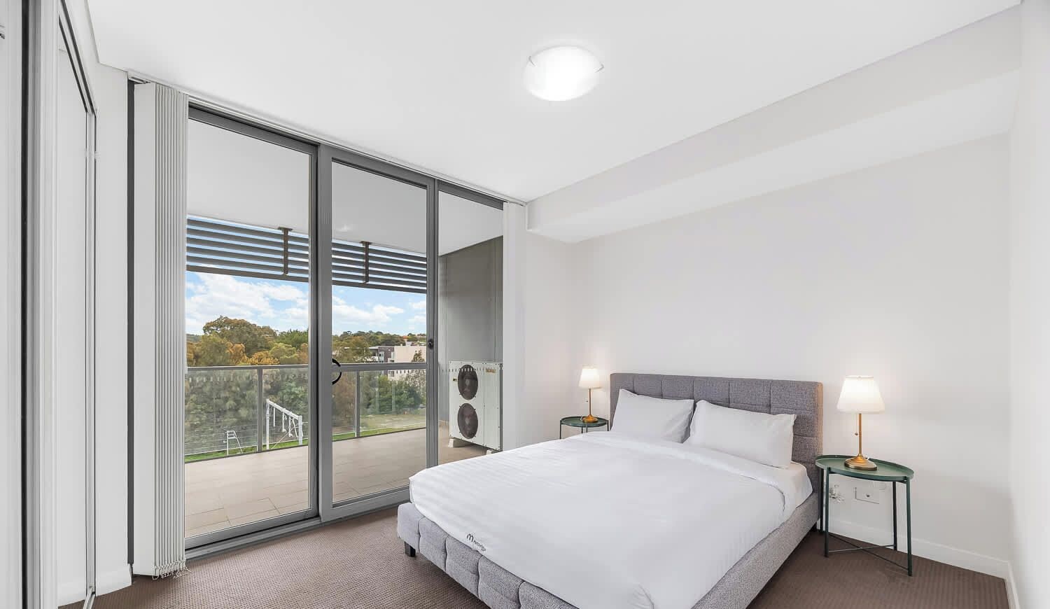 Une chambre d’hôtel moderne avec un grand lit, deux tables de chevet, une porte-fenêtre coulissante donnant sur un balcon et un luminaire au plafond.
