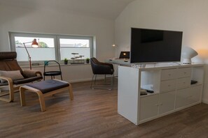 Ein modernes Wohnzimmer mit Flachbildfernseher, einer Couch, einem Sessel und einem kleinen Schreibtisch mit Schubladen.