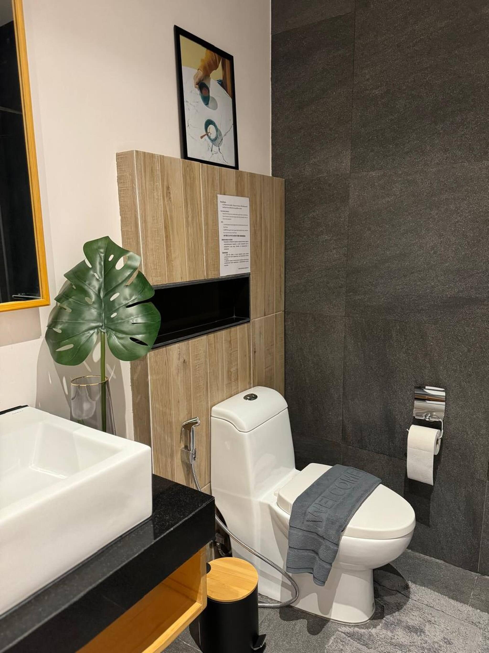 A modern bathroom with a white toilet, a sink, and a shower. There is a framed picture on the wall and a plant on the counter.