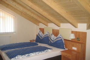 Ein Schlafzimmer mit Holzbalkendecke, einem Bett mit blau-weißer Bettwäsche, einem Nachttisch mit Lampe und einem Fenster mit Jalousien.