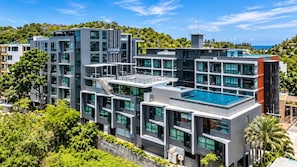 A modern multi-story hotel with a rooftop pool, surrounded by lush greenery and a clear view of the sea. - Sunrise Studio, Nai Harn, Desk, Rooftop Pool (Rawai)