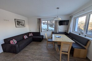 Ein modernes Wohnzimmer mit einem dunklen Sofa, einem Holzespeisetisch und Blick auf eine verschneite Landschaft durch große Fenster.