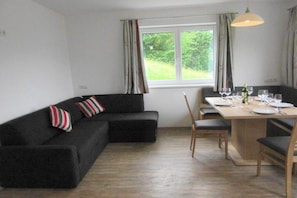 Ein modernes Wohnzimmer mit einem schwarzen Ecksofa, einem Esstisch für vier Personen und einem Fenster mit Vorhängen.
