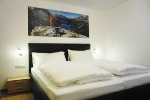 Ein ordentlich bezogenes Bett mit weißen Leinen und Kissen, ein Nachttisch aus Holz mit einer Schublade und ein Wandbild von einer Berglandschaft.