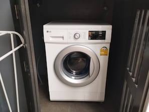 A white LG front-loading washing machine in a laundry room with a metal rack to the left. - Nai Harn Studio, Steps to Everything, Pool, Gym (Rawai)