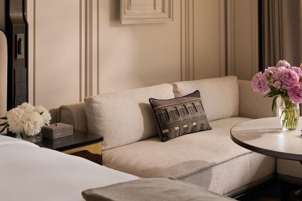 A hotel room with a sofa, a small table with a vase of flowers, and a bedside table with a lamp.