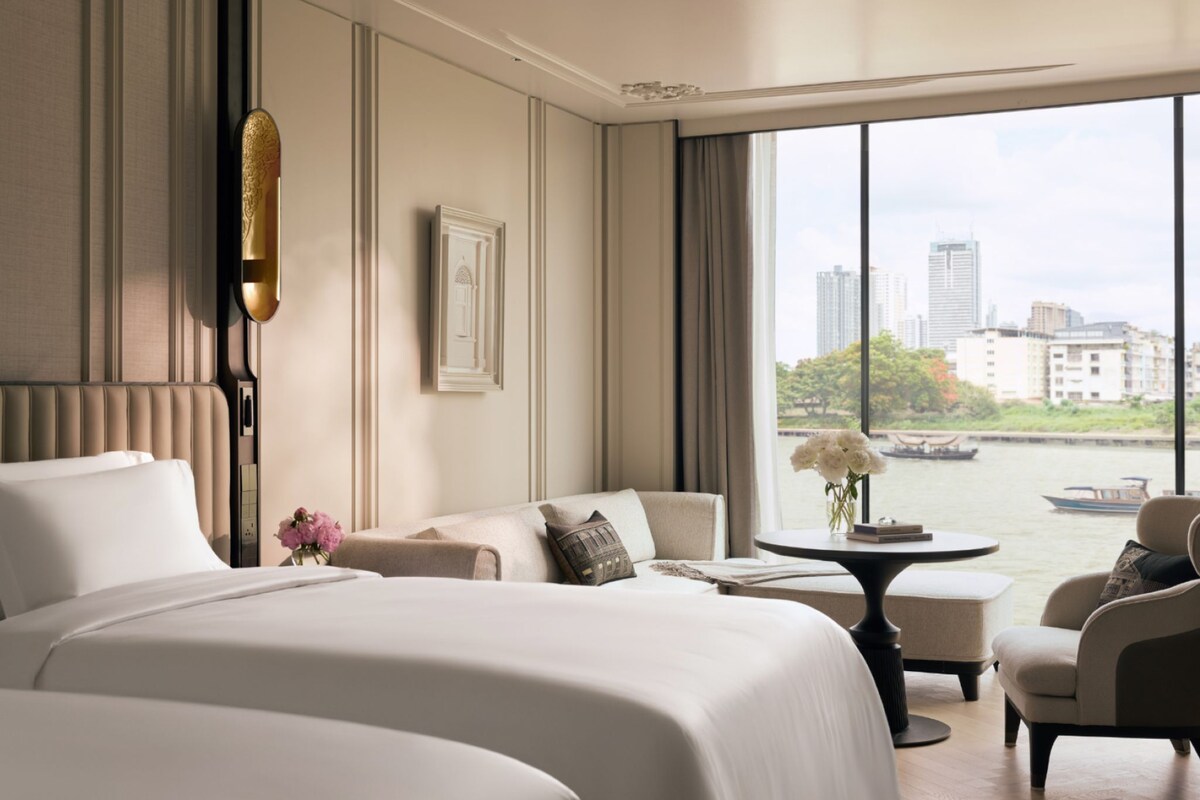 A hotel room with a large bed, a sofa, a small table, and a view of a cityscape through a large window.