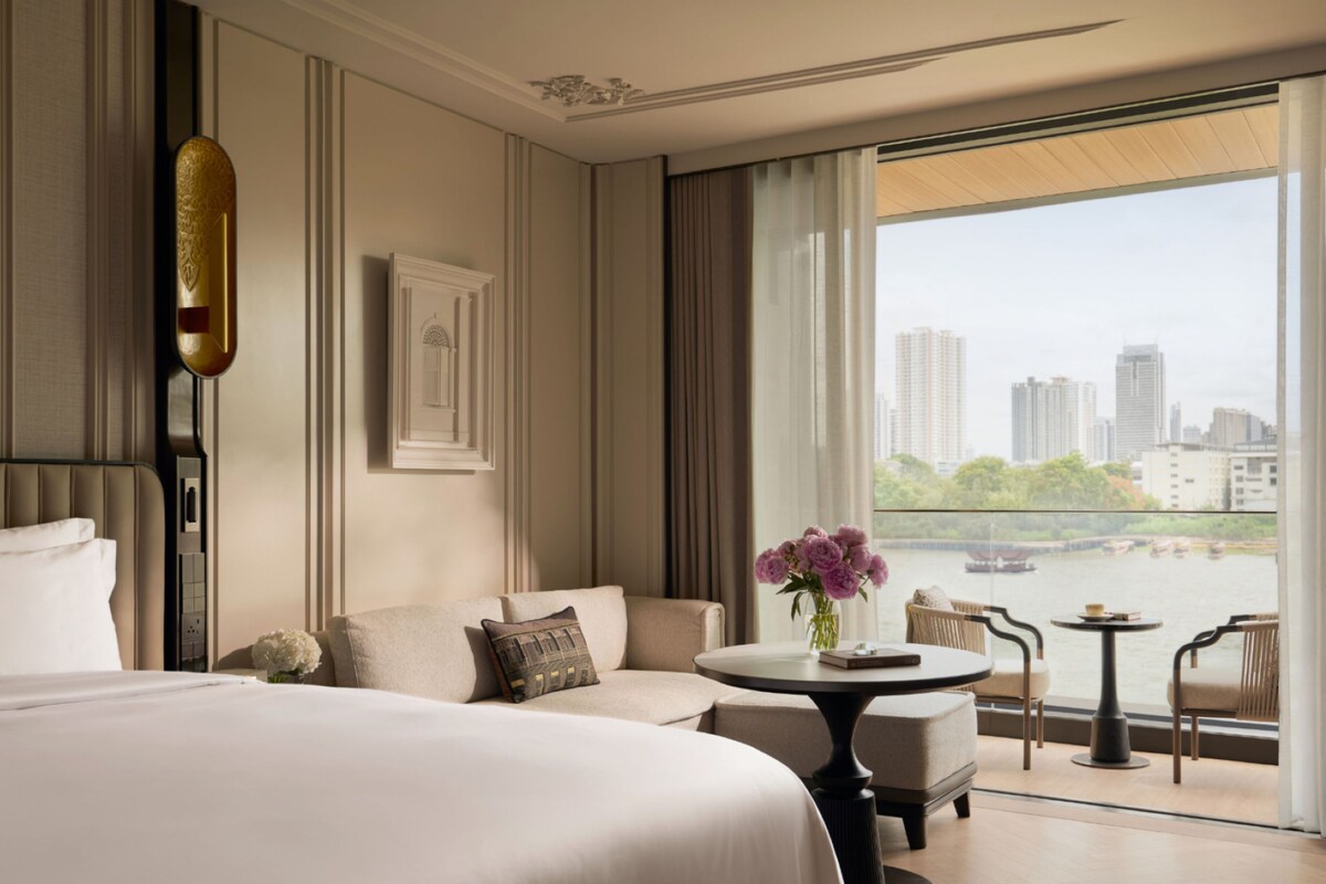 A hotel room with a bed, a sofa, a small table, and a view of a cityscape through large windows.