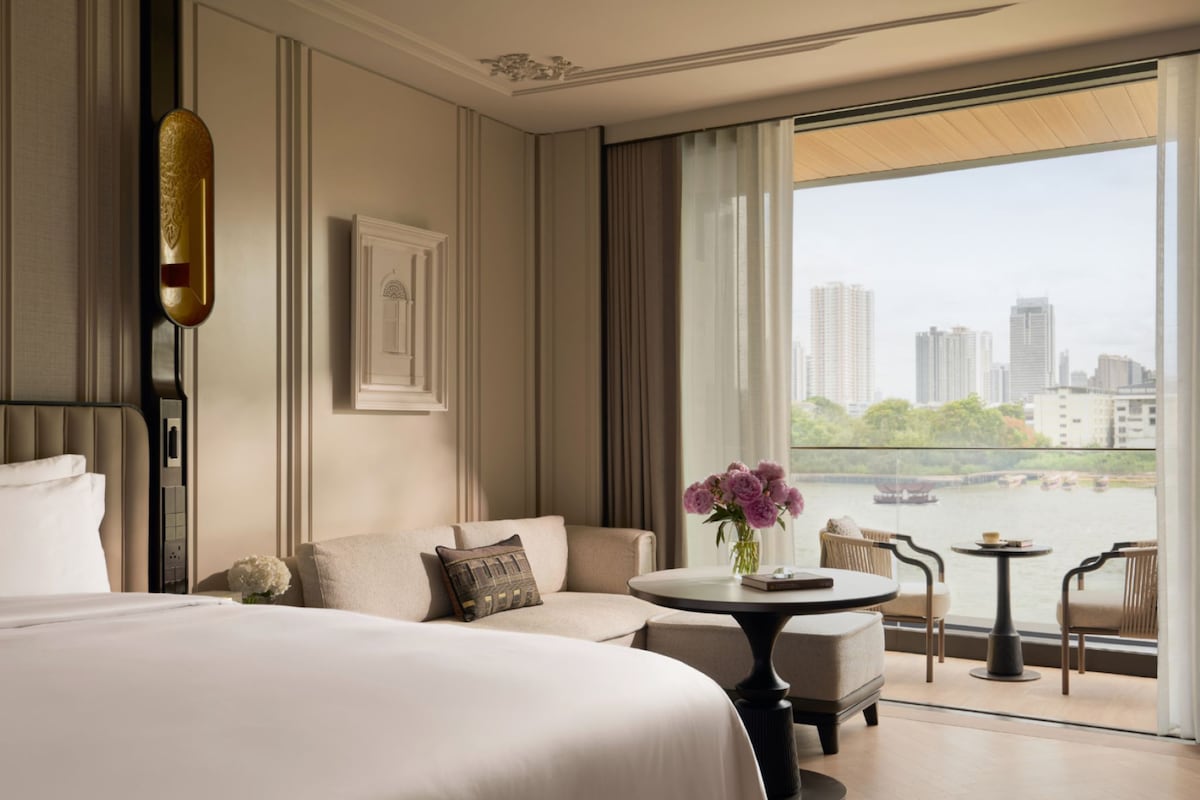 A hotel room with a large bed, a sofa, a small table with a vase of flowers, and a view of a cityscape through a large window.