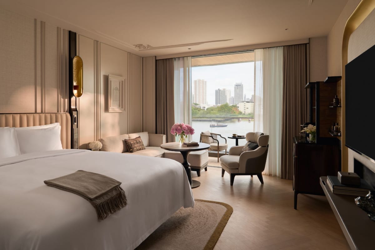 A hotel room with a large bed, a sitting area with a table and chairs, a TV, and a view of the city.
