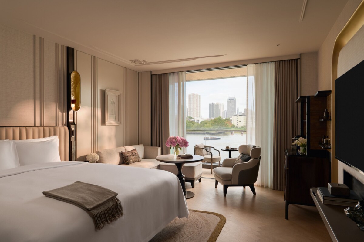 A hotel room with a large bed, a sitting area with a table and chairs, a television, and a view of a cityscape through large windows.