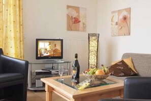 Un salon avec une télévision, une table basse en verre, une bouteille de vin et un panier de fruits.