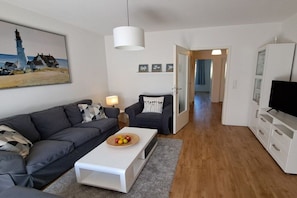 A living room with a sofa, a coffee table with fruit, a television, and a painting of a lighthouse. - Holiday apartment with 2 bedrooms (Ostseebad Boltenhagen)