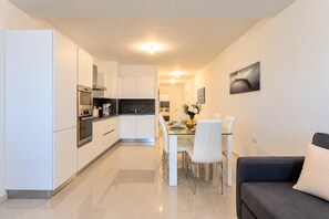 A modern kitchen with white cabinets, a dining area with a glass table, and a dark-colored sofa. - Bright 2BR Flat in St. Julian's - Balcony & Views (St. Julian's)
