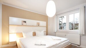 Ein Schlafzimmer mit einem großen Bett, einem Nachttisch, einem Fenster mit Vorhängen und einem an der Wand befestigten Regal mit Bildern.