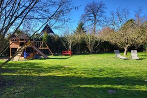 Casinha de madeira com escorrega e parede de escalada, um banco vermelho e duas cadeiras de descanso em um gramado com árvores.