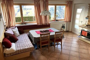 Une pièce confortable avec un canapé, une table à manger et des chaises. On y trouve une fenêtre avec des rideaux, un radiateur et un coin cuisine équipé d’une cuisinière.