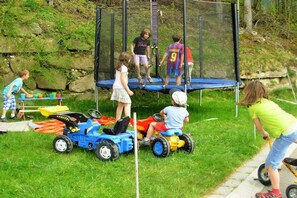 Des enfants jouent sur un trampoline et conduisent des véhicules jouets dans un espace extérieur herbeux.