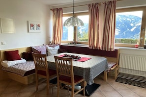 Un coin repas accueillant avec une table, des chaises et un banc. Une fenêtre avec des rideaux offre une vue sur les montagnes.