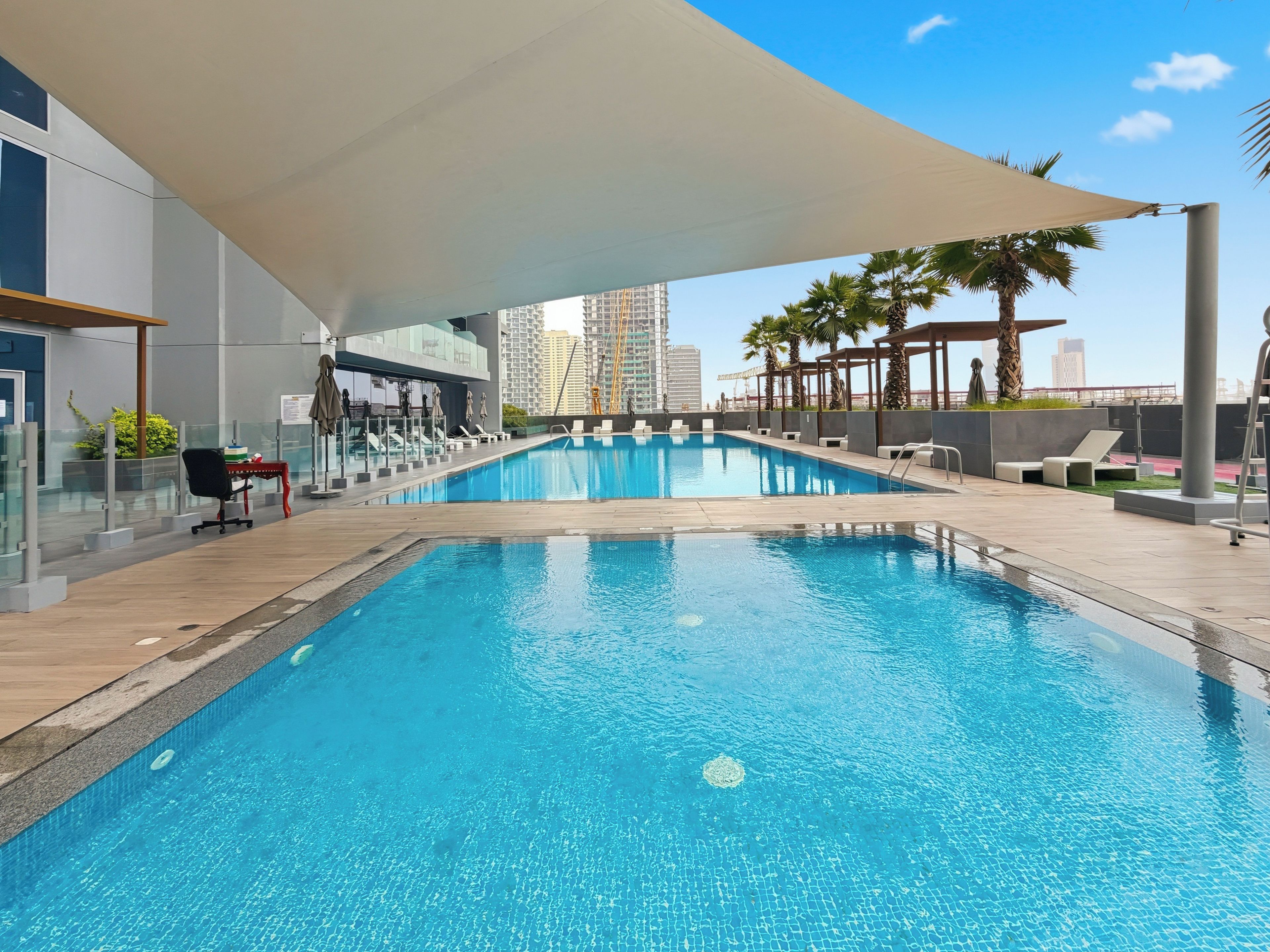 A swimming pool with a sunshade, lounge chairs, and a view of tall buildings in the background.