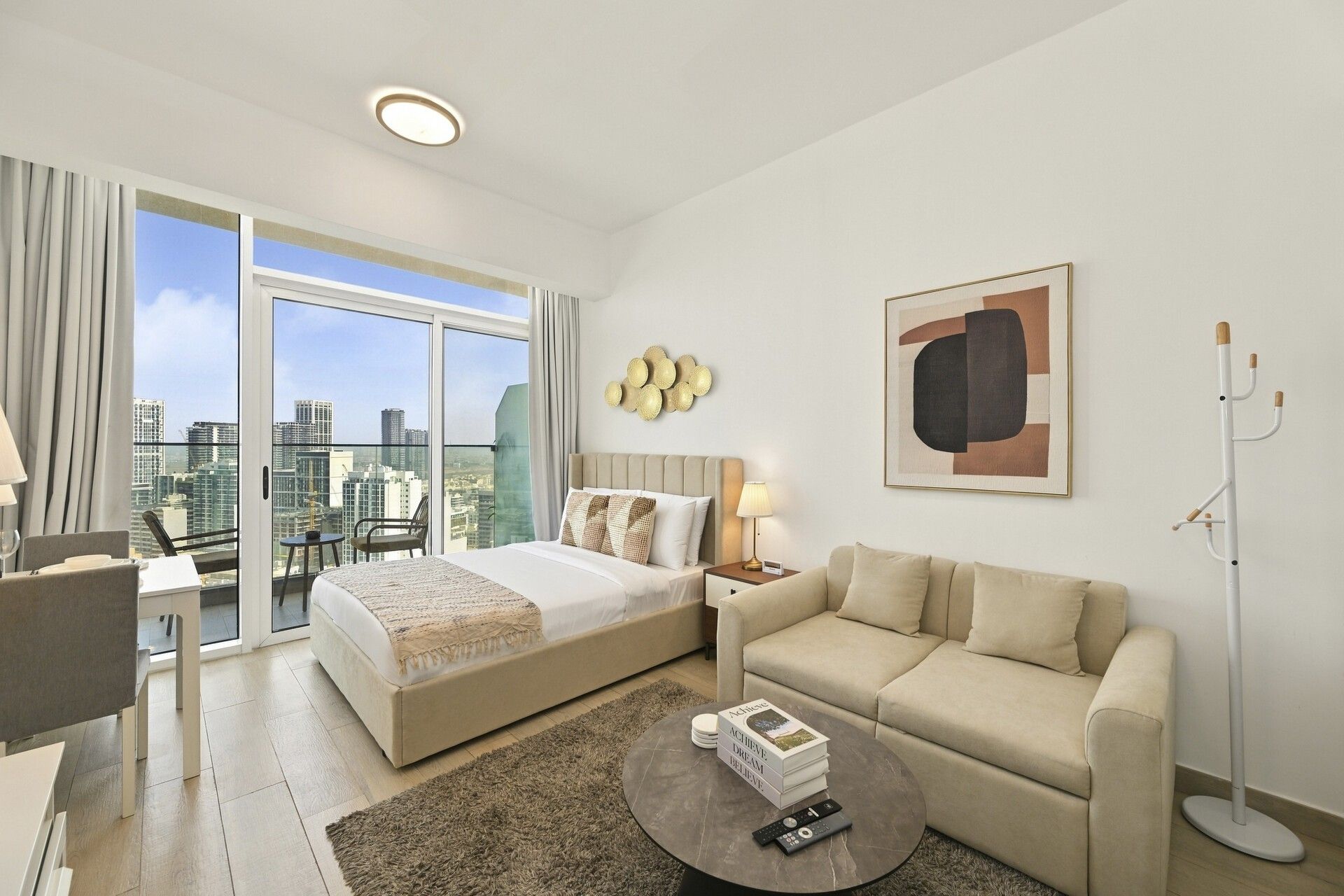 A hotel room with a bed, a sofa, a coffee table, a desk, and a chair. There is a large window with a city view.