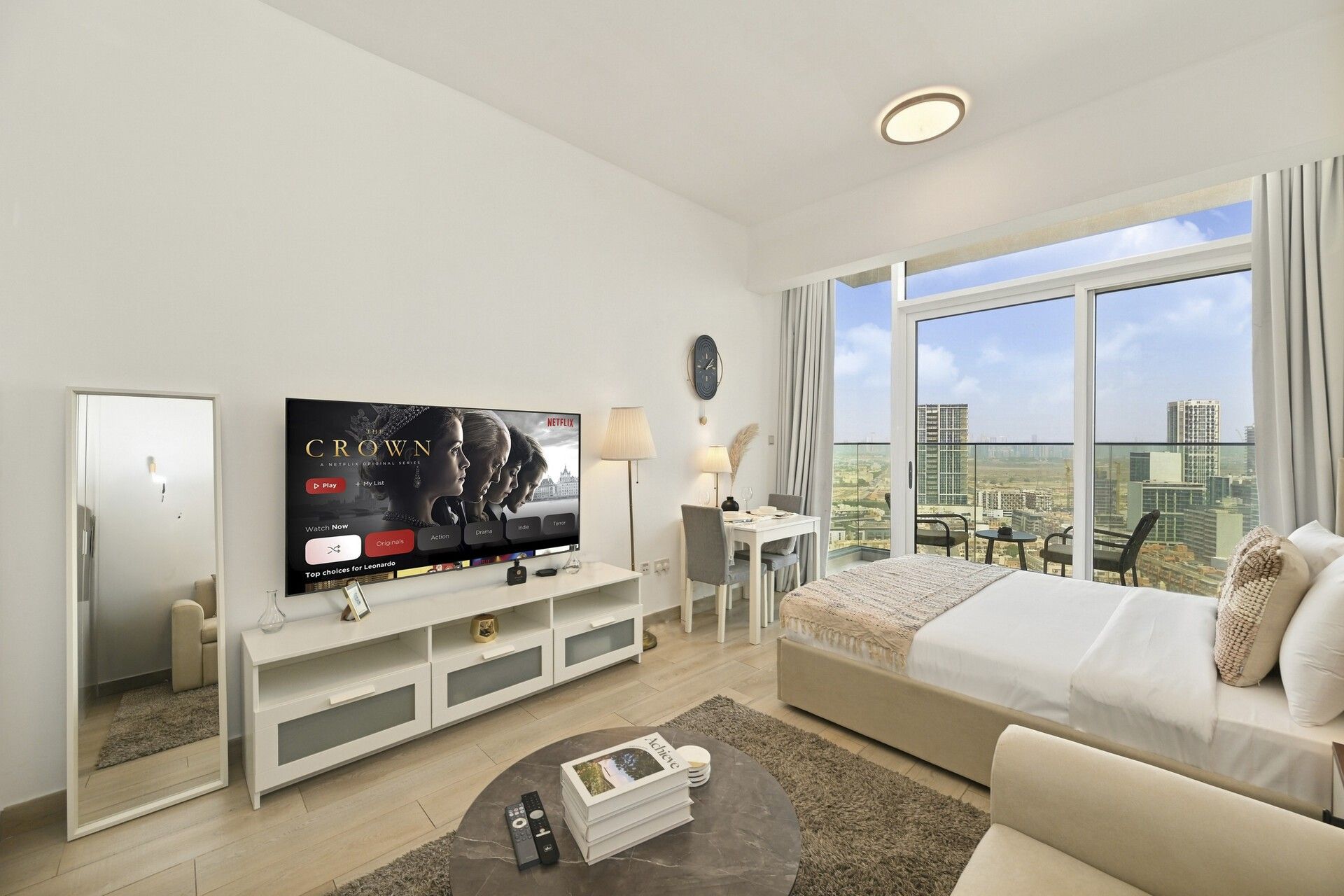 A modern hotel room with a large TV, a bed, a seating area, and a balcony with a city view.