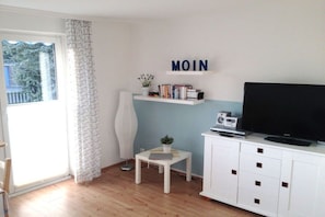 Ein Zimmer mit Fernseher, einem kleinen Tisch mit Pflanze, einem Regal mit Büchern und einem Fenster mit Vorhang.