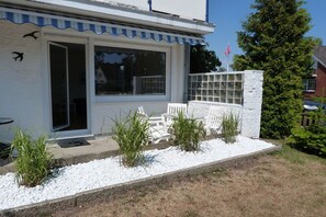 Ein Haus mit weißer Fassade, einem großen Fenster und einer kleinen Veranda mit weißem Zaun und Pflanzen.