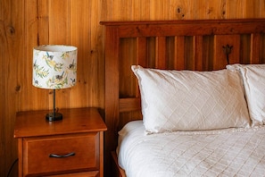 Ein hölzernes Bett mit weißer, gesteppter Bettwäsche, ein Nachttisch aus Holz mit einer Lampe mit Vogelmuster und einer Schublade.